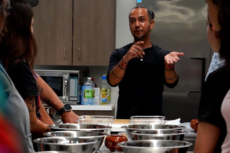 Man standing at foot of long table talking as others listen about cooking.
